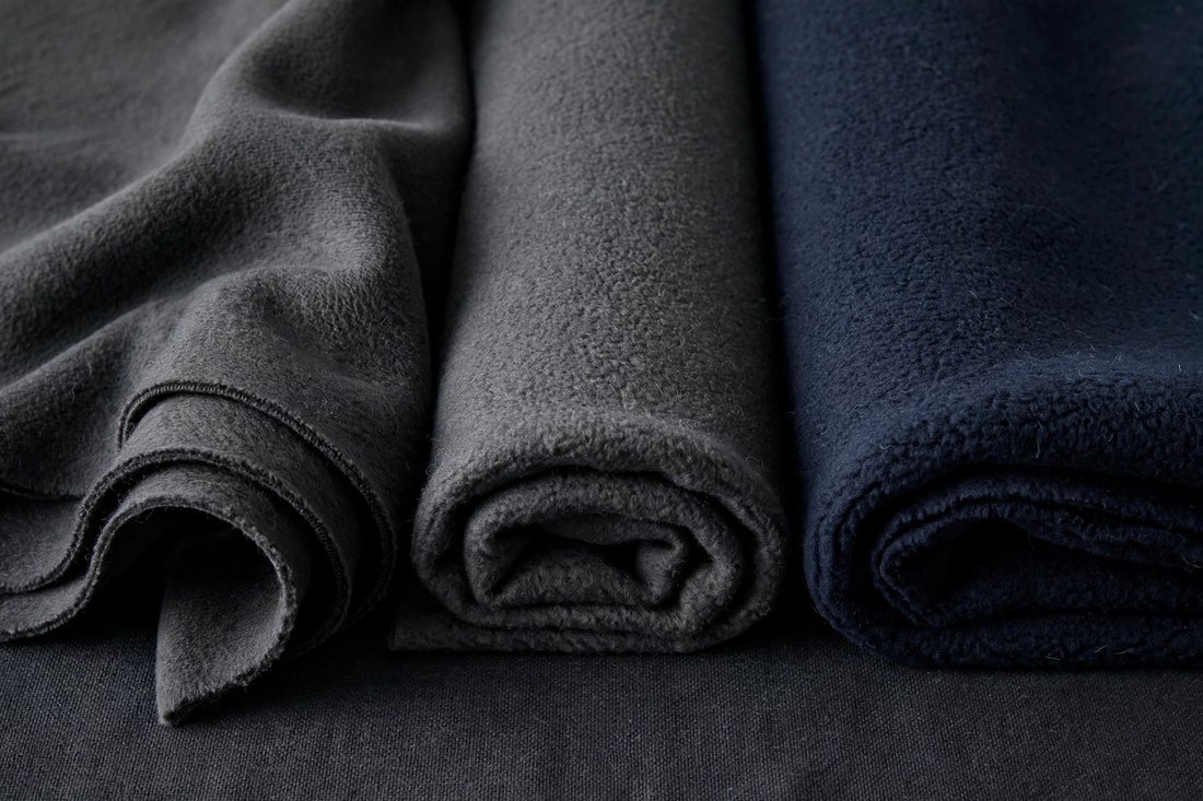 Three folded pieces of fleece in charcoal, grey and navy laid side by side, each turned back to show the brushed inner face and its thickness